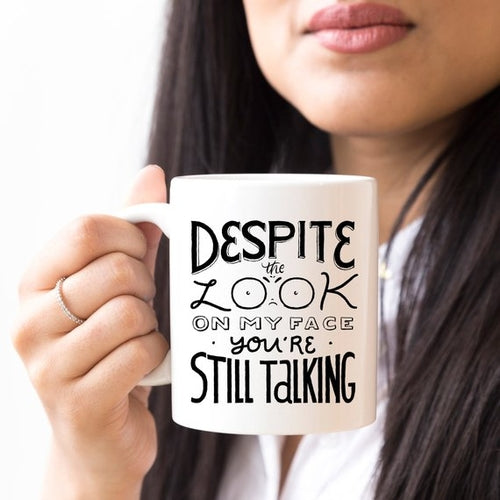 Tazza nera Sajishop con scritta "Despite The Look On My Face, You're Still Talking" in caratteri bianchi, manico resistente.