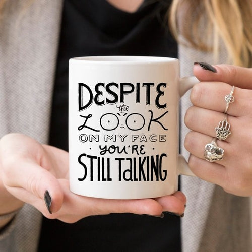 Tazza nero Sajishop con manico, 11 oz, con testo "Despite The Look On My Face, You're Still Talking" su superficie bianca.